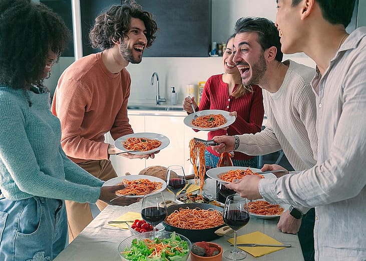 Five people gathered around a table standing with plates full of spaghetti.