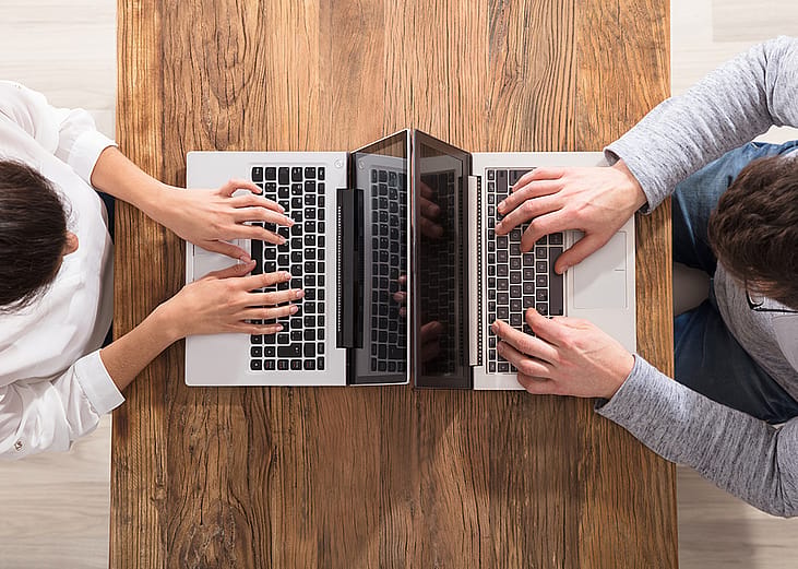 An overhead view of two people using laptops and sitting across each other on a table.