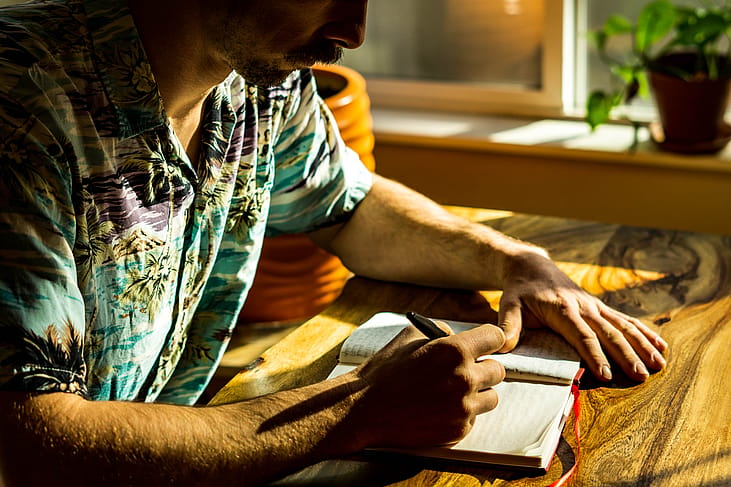 Man wearing a Hawaiian-style shirt sitting next to the window with light coming in, writing in a journal. 