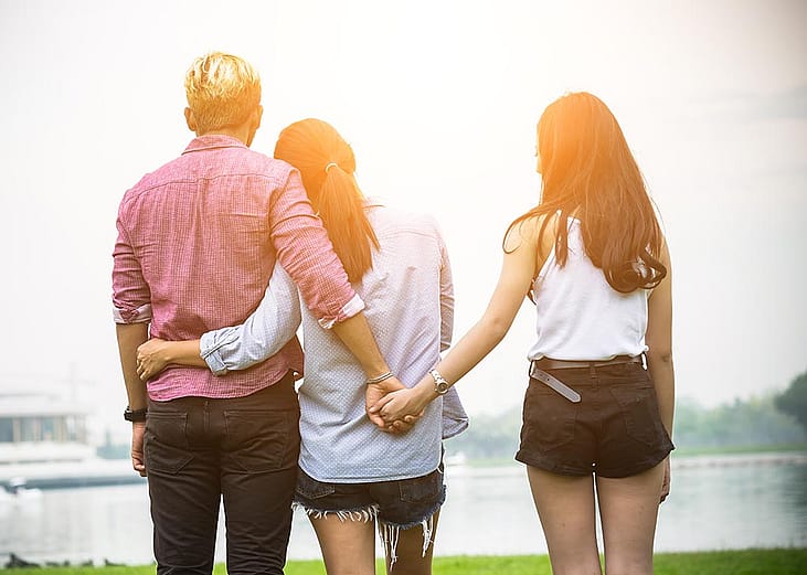 Couple with their back to the camera is looking at a scenic view with their arms around each other, but man is secretly holding hands with another person behind the woman's back.