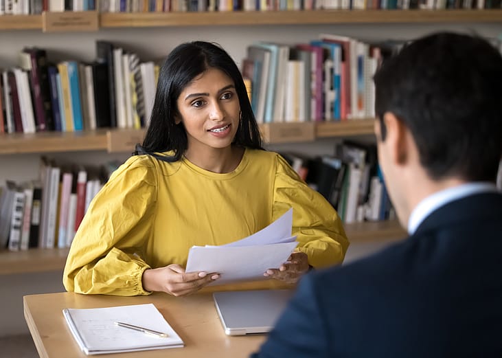 A woman behind a table holding papers and talking to a man sitting on the other side.