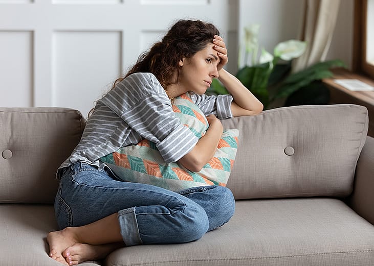 A stressed young woman, sitting at a sofa and cuddling a pillow while lost in thought.