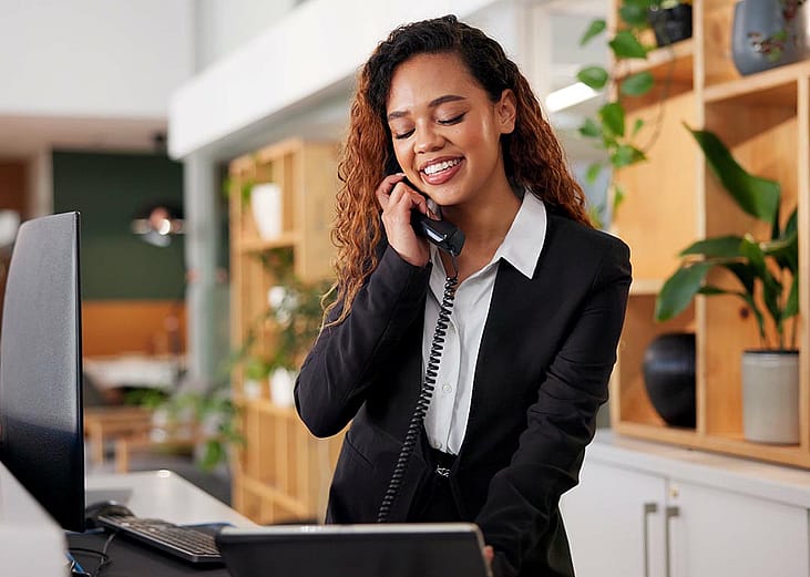 Customer service concierge answering phone standing behind reception desk looking at computer screen with modern decor and houseplants behind her.