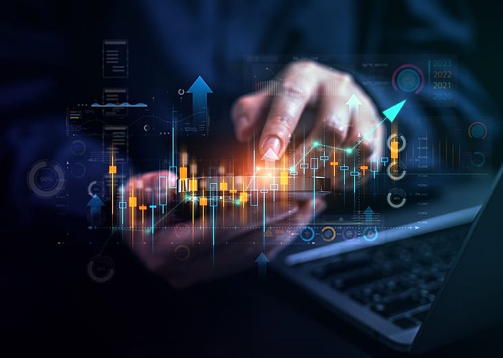 Stock market trading graphics illustrated over a businessperson using a phone and laptop.