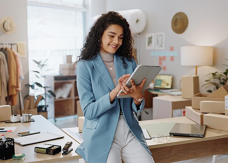 Fashion designer wearing powder-blue oversized blazer and white pants uses a tablet and tablet working logistics or small business managementwith office, shipping and clothing rack behind her.