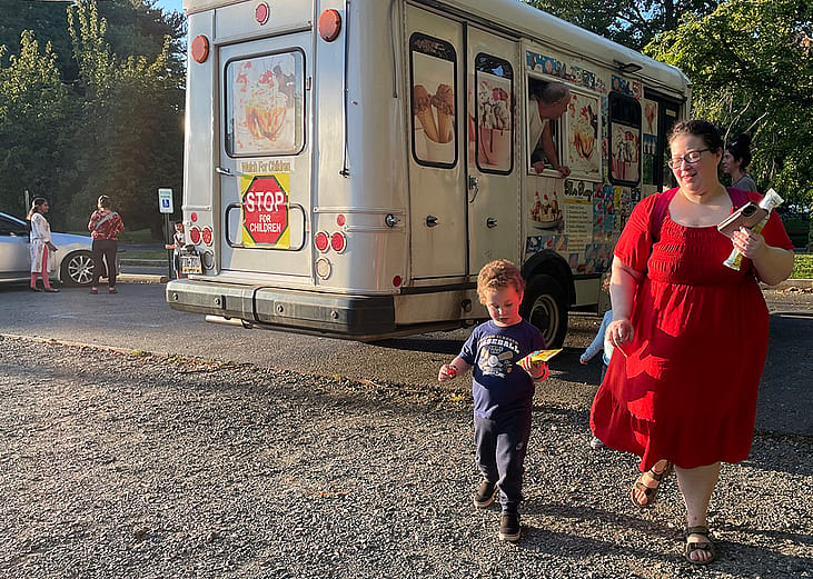 Kimberly Halevy wearing a crimson red midi dress with mid-length sleeves walks with her son Joshua beside her, enjoy treats from an ice cream truck visible behind them, after a recent session with a special instructor.