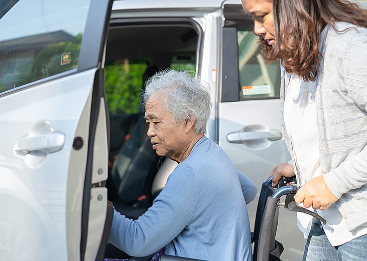 An elderly senior woman in a wheelchair being assisted by her caregiver getting in a car.