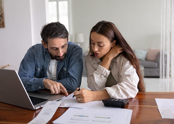 Serious young to middle aged couple discussing expenses at table with papers, a laptop and a calculator.
