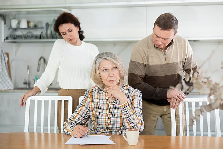 A senior woman signing documents with adult children behind her.
