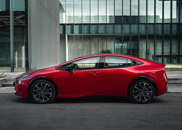 A red 2024 Toyota Prius Prime parked in front of a building on a city street.