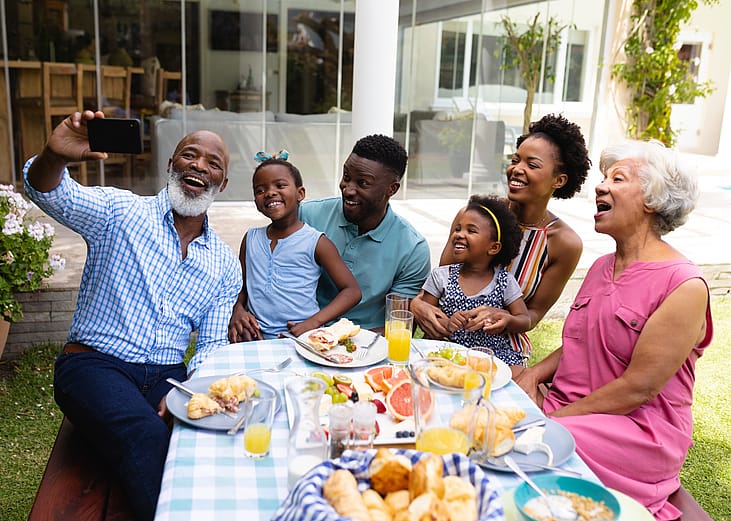 An older man smiling while taking selfie with his family at an outdoor dining table during brunch. 