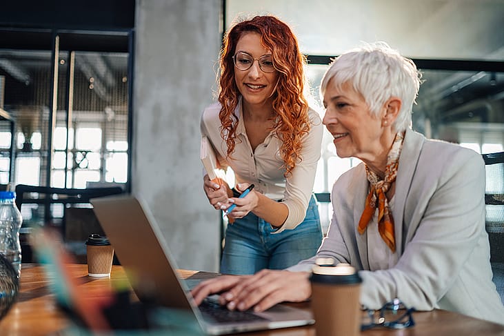 A senior employee working with a younger colleague on a task in an office.
