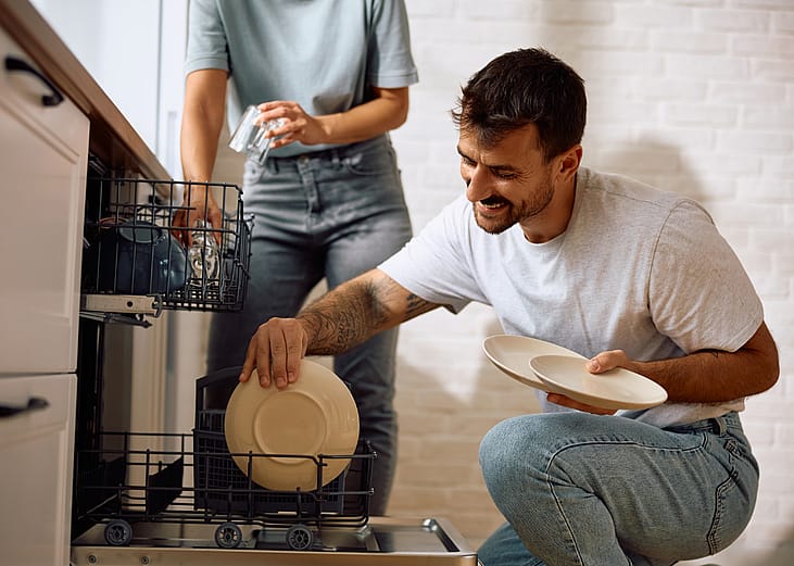 Happy man taking dishes out of the dishwasher while someone stands in the background.