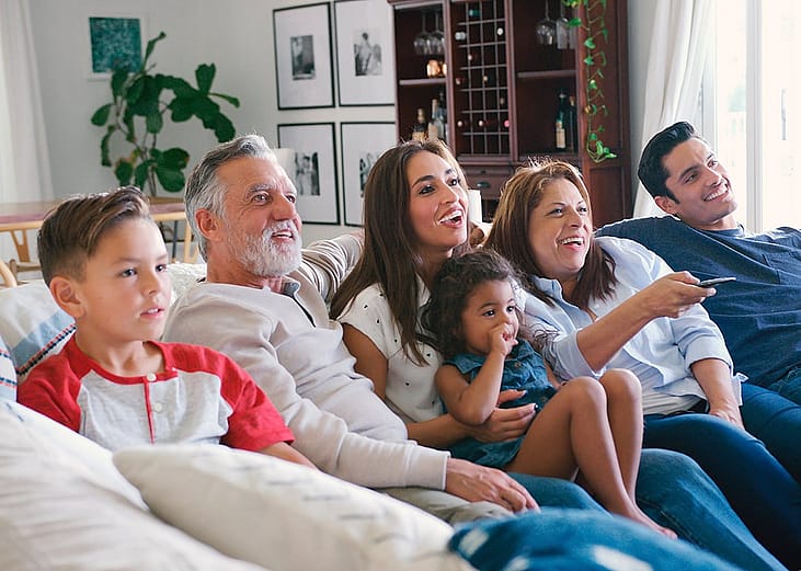 A multi-generational family sitting together on a sofa and watching televisions.