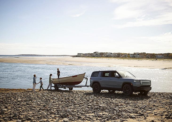 Rivian R1S towing a boat, parked near a body of water on a rocky dirt beach. A family of four plays near the boat.