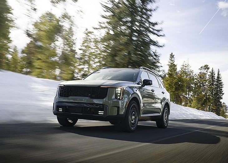 Low-angle view of earth-toned green 2026 Kita Sorento driving on mountain road with motion-blurred snow and pine trees in the background.