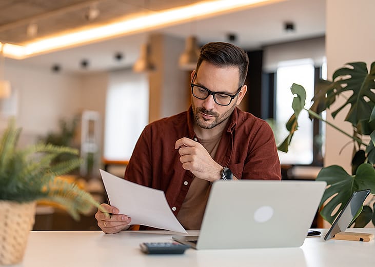 A person looking at a paper and using a laptop in a home office. 