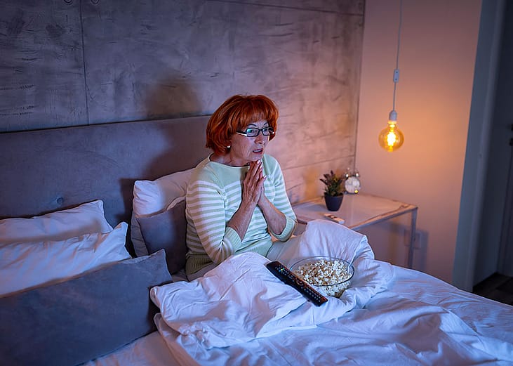 A senior woman watching a scary movie on the TV from bed.