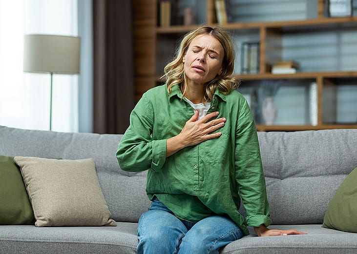 A woman sitting on a sofa holding her chest due to pain.