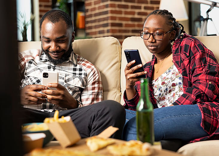 A black couple relaxing on a sofa at home and individually using their smartphones.