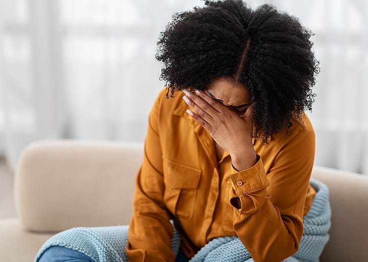 A young black woman sitting on a sofa holding her due to stress.