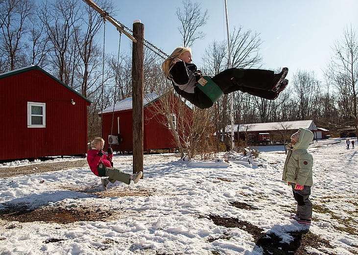 Two children on a swingset while another child watches on a snowy day outside Nature's Gift Microschool.