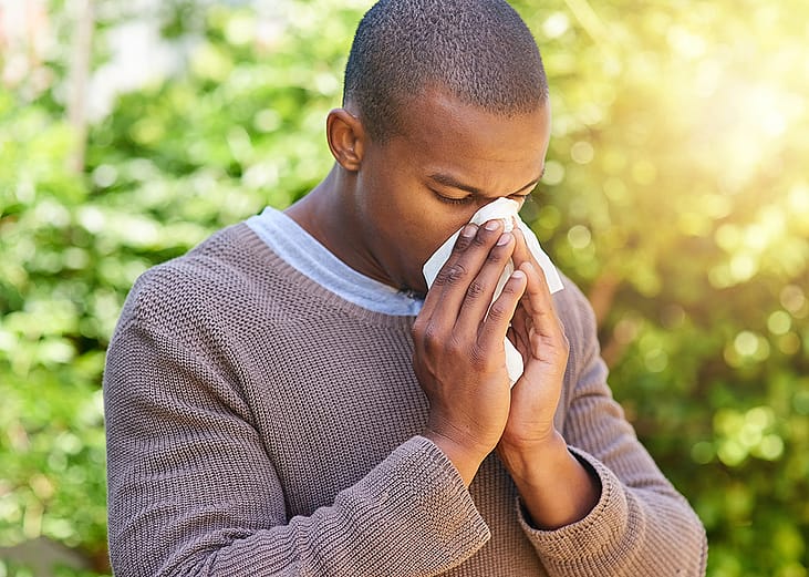 A young black man blowing nose outdoors due to allergy.