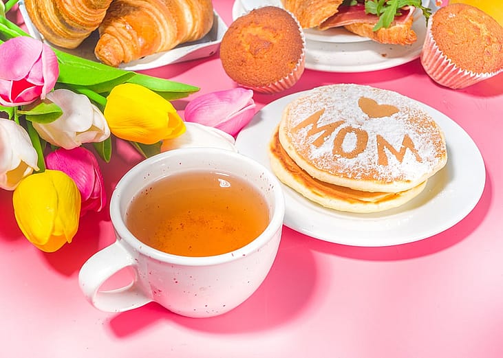 A pink-themed Mother's Day brunch meal featuring a bouquet of flowers, a cup of tea, and pancakes.