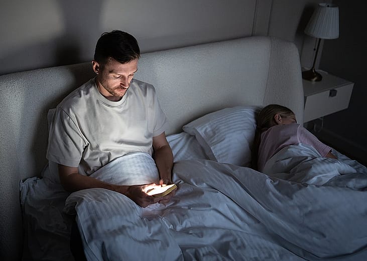 A man awake late at night and scrolling through his phone beside his asleep wife.