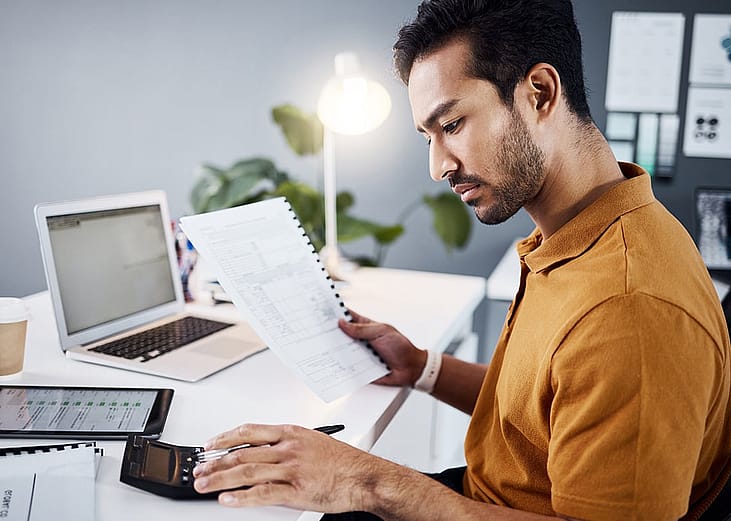 A finance analyst reviewing financial documents using a calculator and a work laptop in an office.