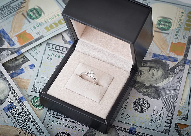 A diamond engagement ring placed above U.S. dollar bills.