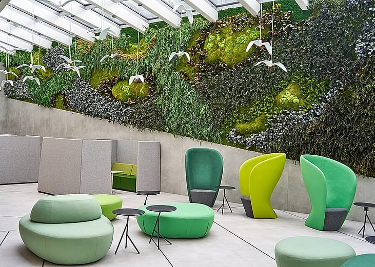A modern office interior featuring a large biophilic living wall covered in various mosses, ferns, and plants spanning the back wall beneath a glass skylight ceiling with white beams. The space includes contemporary furniture in shades of green and lime.