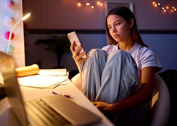 A young woman sitting at a desk in her bedroom and browsing her phone at night.