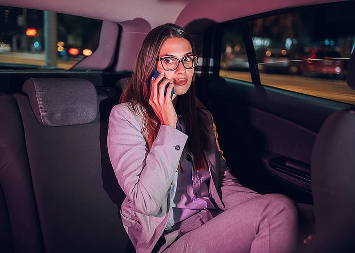A young businesswoman in the backseat of a car late at night and talking on the phone.