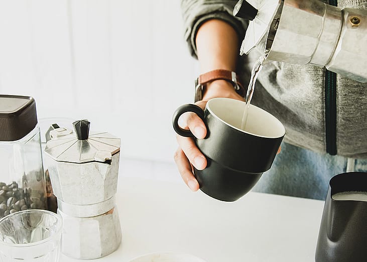 A person pouring hot water from a moka put to a cup before brewing coffee beans.