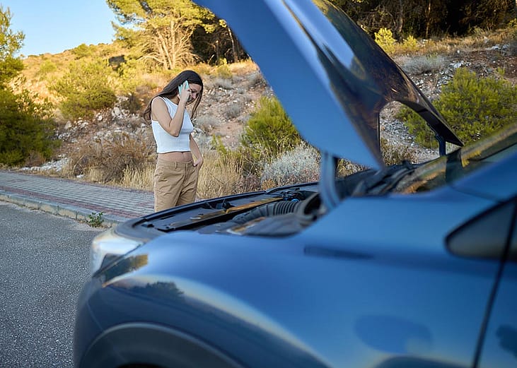 A girl standing beside her broken car on the side of a road.