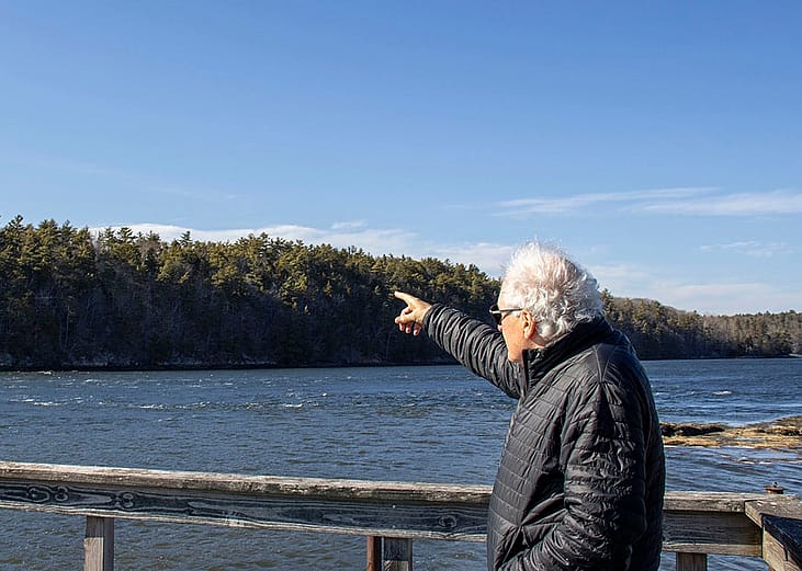 Protect Wiscasset co-founder Richard Davis facing away from the camera wearing a black jacket and his white air blown by the wind, points to the undeveloped land across a waterway where the prospect of a $5 billion data center emerged.