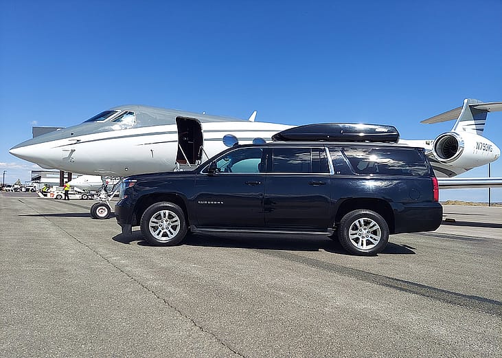 A black Chevrolet Suburban car parked alongside a private air jet in an open ground.