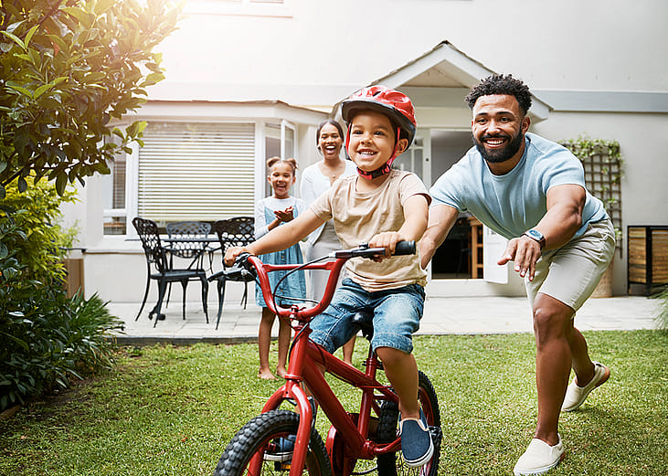 A young boy learning how to ride a bicycle being cheered on by his family.