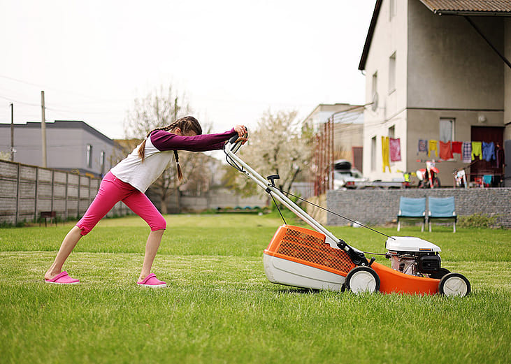 A teenager pushing an electric lawnmower in their backyard during a summer holiday.