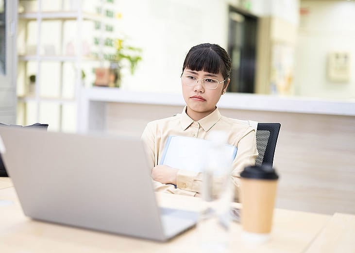 An employee thinking on a work-related matter while looking at a laptop from her office desk.