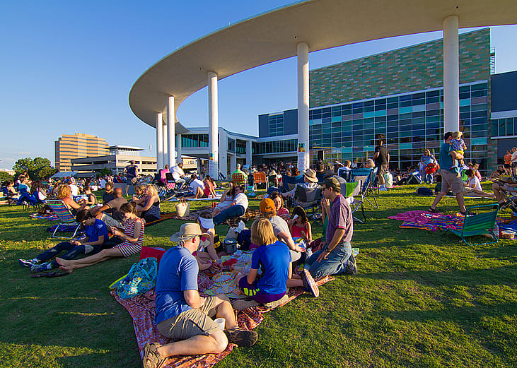 Groups of people sit on grass as they wait for an outdoor concert at the Long Center Lawn in Austin, Texas.