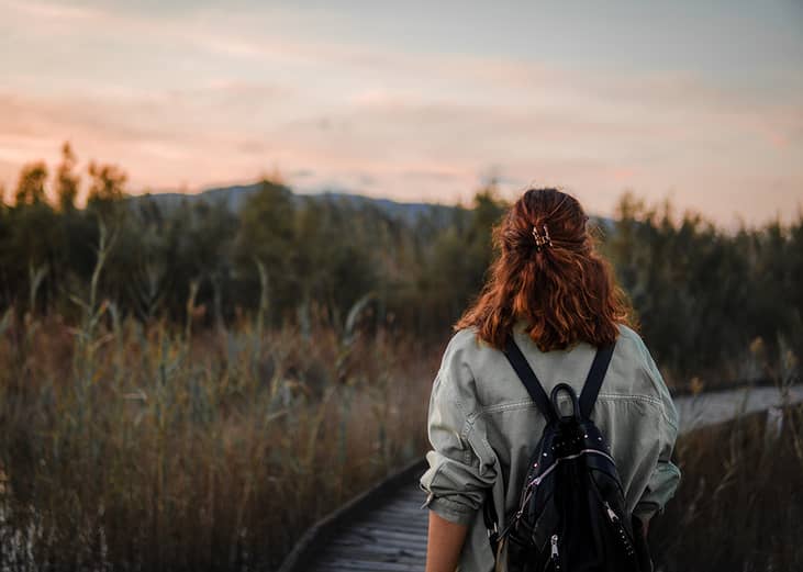 A girl seen from behind with a backpack walking through nature at sunset.