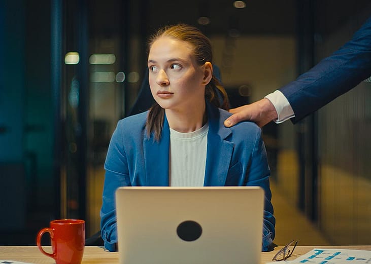 A young business assistant appearing to be uncomfortable as a male colleague places a hand on her shoulder.