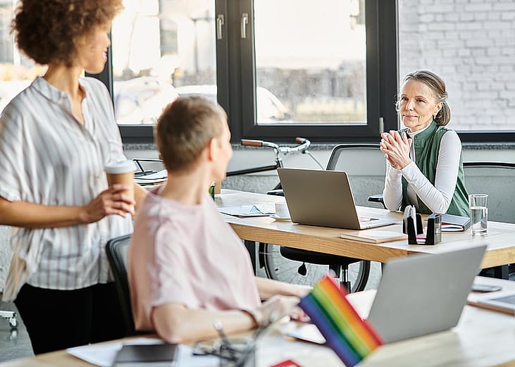 Business partners collaborating on a project and a rainbow flag seen from a desk indicating it as a LGBTQ+ inclusive workplace. 