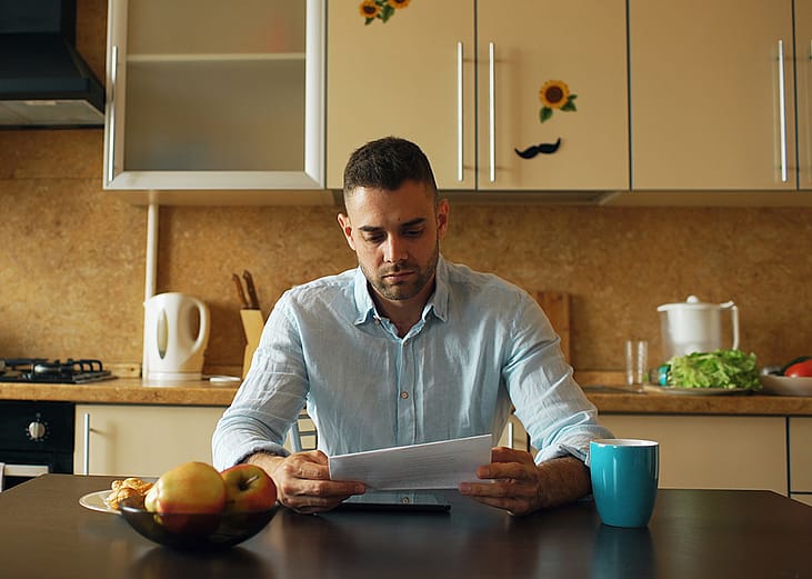 A male homeowner reading an unpaid bill letter in his kitchen.