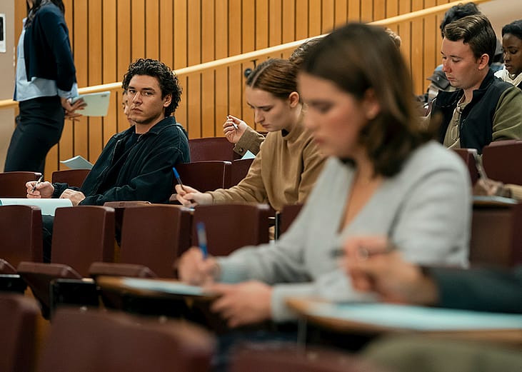 A publicity still from the first season of the TV show Off-Campus shows two people in the same college lecture with the focus on one man watching another woman while seated in the row behind her.