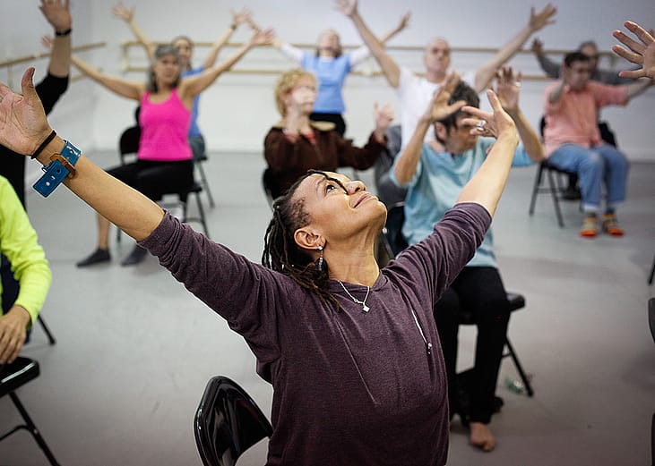 View of upturned face of Black woman with her arms extended, leading a chair dance class with lots of seniors sitting in the background extending their arms in the same motion.