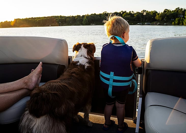 A toddler wearing a life vest and their family dog looking at the waters from a boat. 