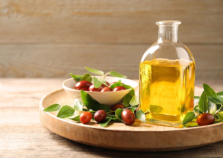 A glass bottle of jojoba oil with leaves and seeds of a shrub in a wooden plate.